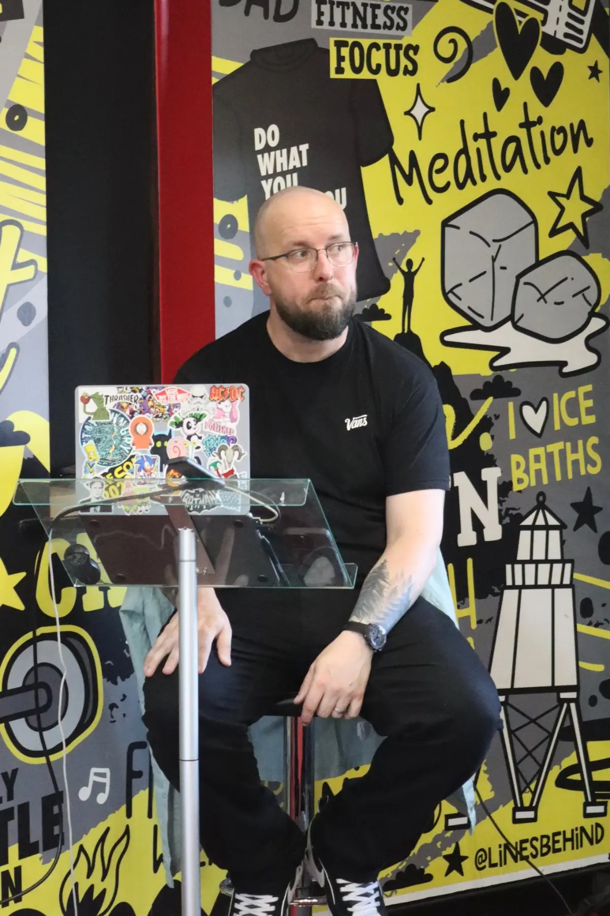 Joel Stone seated at a lectern with a laptop, in front of a hand-drawn mural